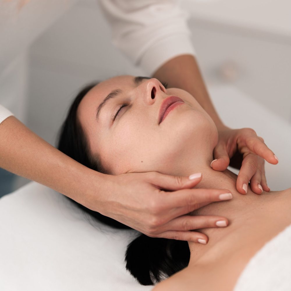 From above of crop unrecognizable therapist massaging neck of female client gently, lying on table with closed eyes during rehabilitation session in modern light spa salon